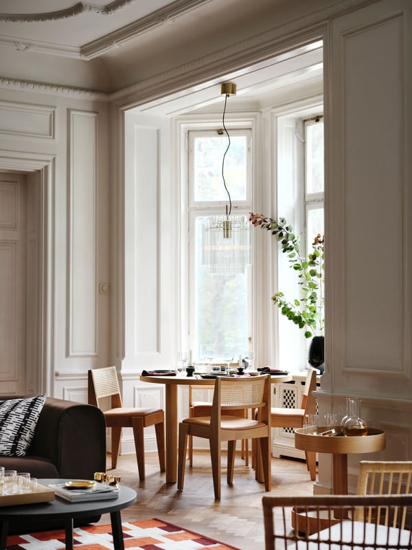 A living room dining set with an oak veneer STOCKHOLM 2025 table and oak and rattan chairs placed by large bay windows.