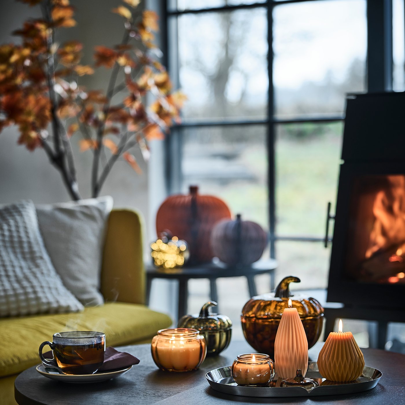 Un rincón acogedor junto a una ventana, con una mesa de centro en la que hay velas perfumadas HÖSTAGILLE encendidas, velas gruesas con forma de calabaza y una taza.