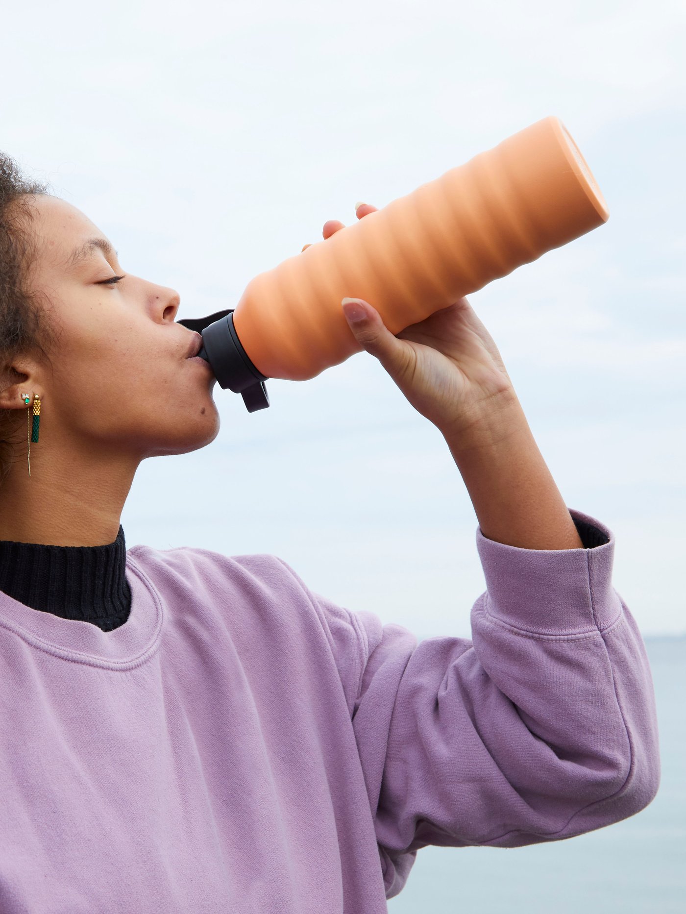 A woman wearing a purple sweatshirt standing by the sea drinking from the orange KÅSEBERGA water bottle with a black top.