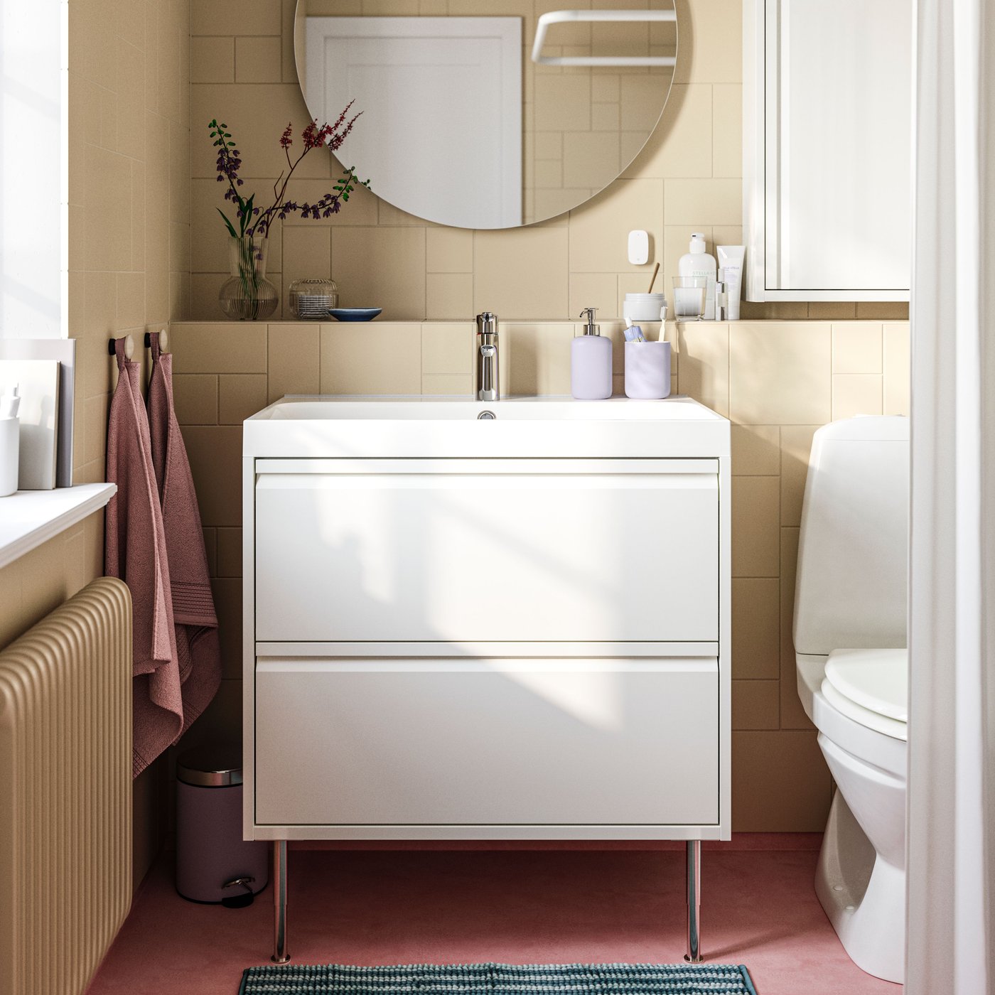 Un meuble lavabo ÄNGSJÖN blanc muni de deux tiroirs et de pieds, avec un miroir FAXÄLVEN le surplombant, dans une salle de bains carrelée en brun clair.