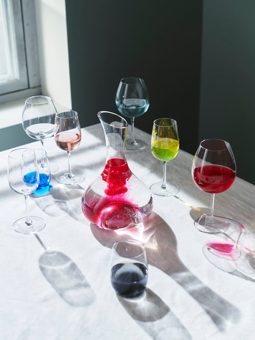 STORSINT glass carafe and drinking glasses with coloured liquid in them on a white tablecloth with strong light and shadow.