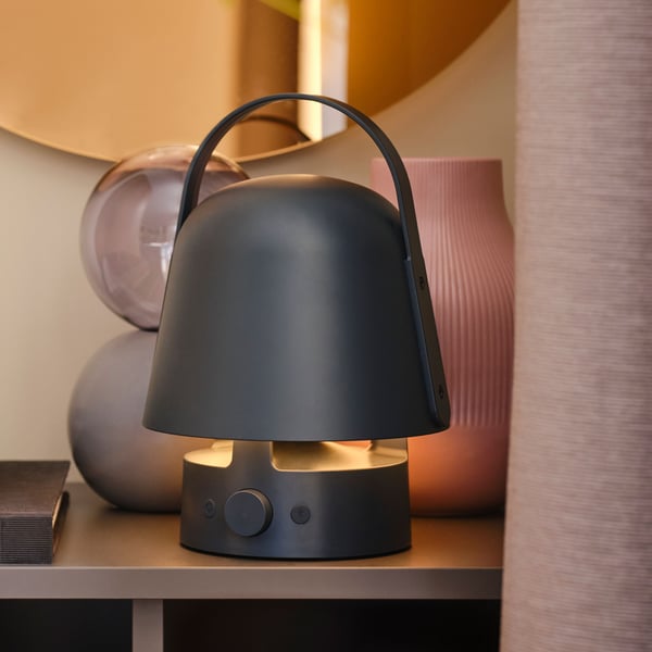 A round, bronze-coloured VÄRMBLIXT LED wall/mirror lamp hangs above a cabinet with decorative items and a speaker lamp on it.
