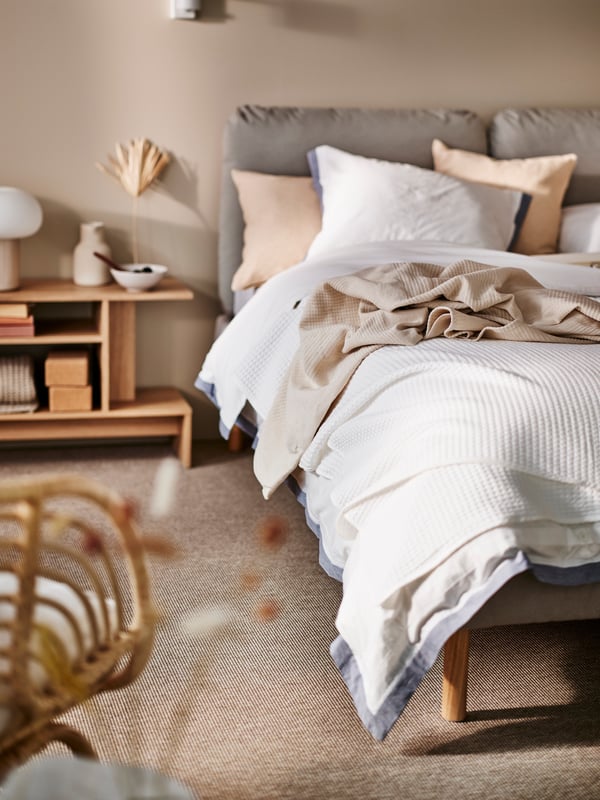 A white SAGESUND upholstered bed in a beige room with white/blue TÅTELSMYGARE bed linen and white VÅRELD bedspread on top.