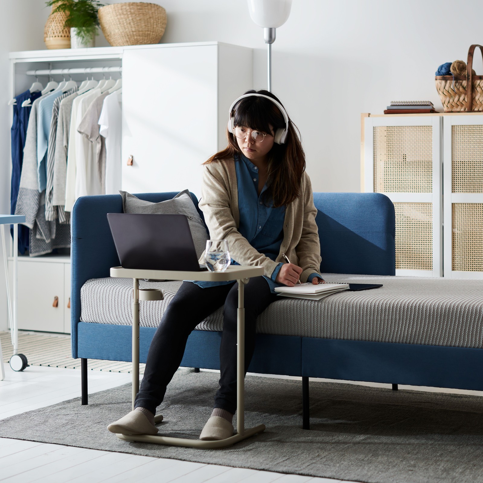A young woman with headphones sits on the side of a BLÅKULLEN bed, focusing on a laptop placed on a BJÖRKÅSEN laptop stand.