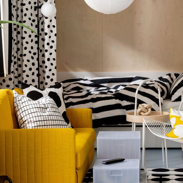 A FRIDHULT sofa-bed stands in a living room area near a white KALLAX shelving unit and a bed with striped bed linen.