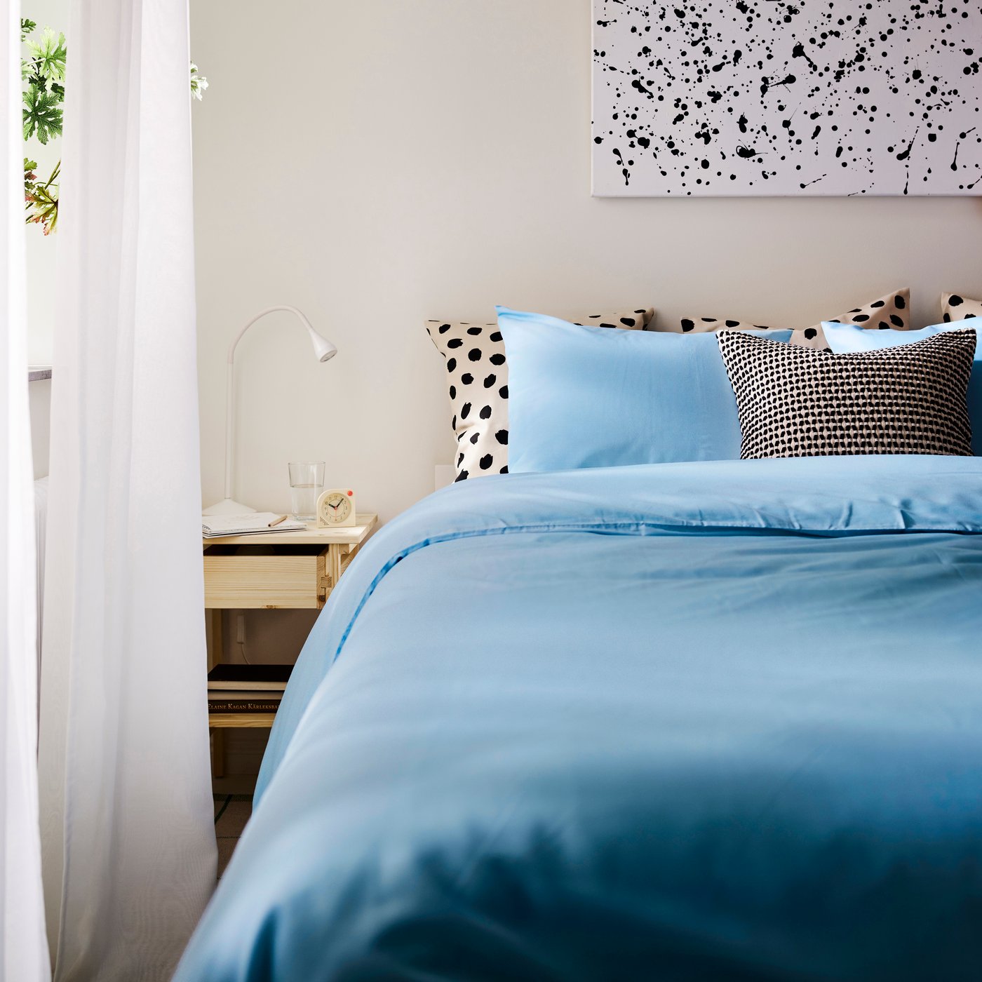 Ein beige‑schwarzer KUSTFLY Kissenbezug im Schlafzimmer. Auf einem Bett liegt eine hellblaue PILTANDVINGE Bettdecke mit passendem Kopfkissen.