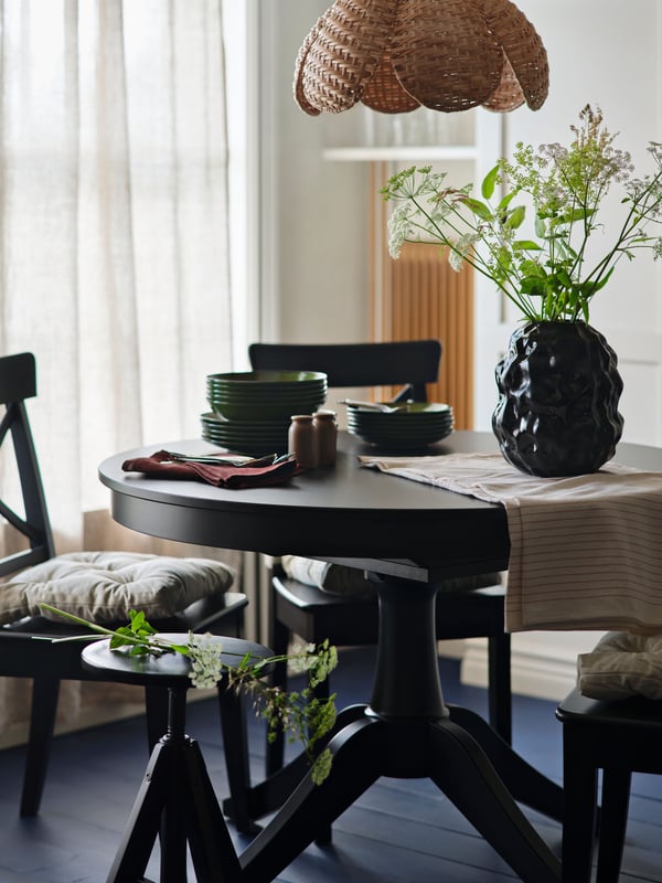 In einem lichtdurchfluteten Esszimmer ist ein schwarzer ROSENTORP Ausziehtisch mit Stühlen, Geschirr und einer Blumenvase gedeckt.