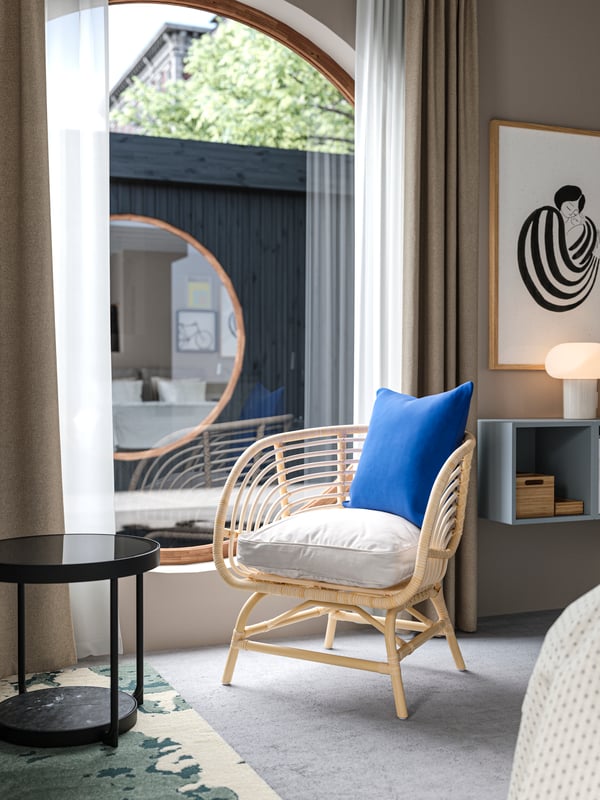 A houseboat hotel room with a BUSKBO rattan armchair and a cushion with a blue cushion cover next to a large, round window.