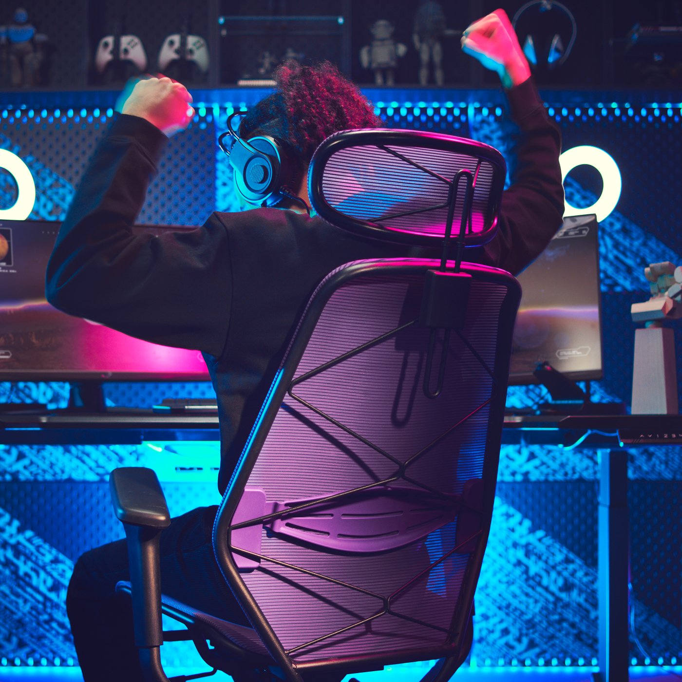 A dark gaming room with a lit-up gaming station. A person, sitting in a gaming chair, is raising the arms victoriously. 