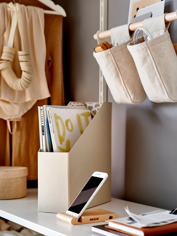 Tablette blanche sur laquelle sont disposés un range-revues TJENA beige et un téléphone sur son support en bambou, sous des sacs en tissu suspendus.