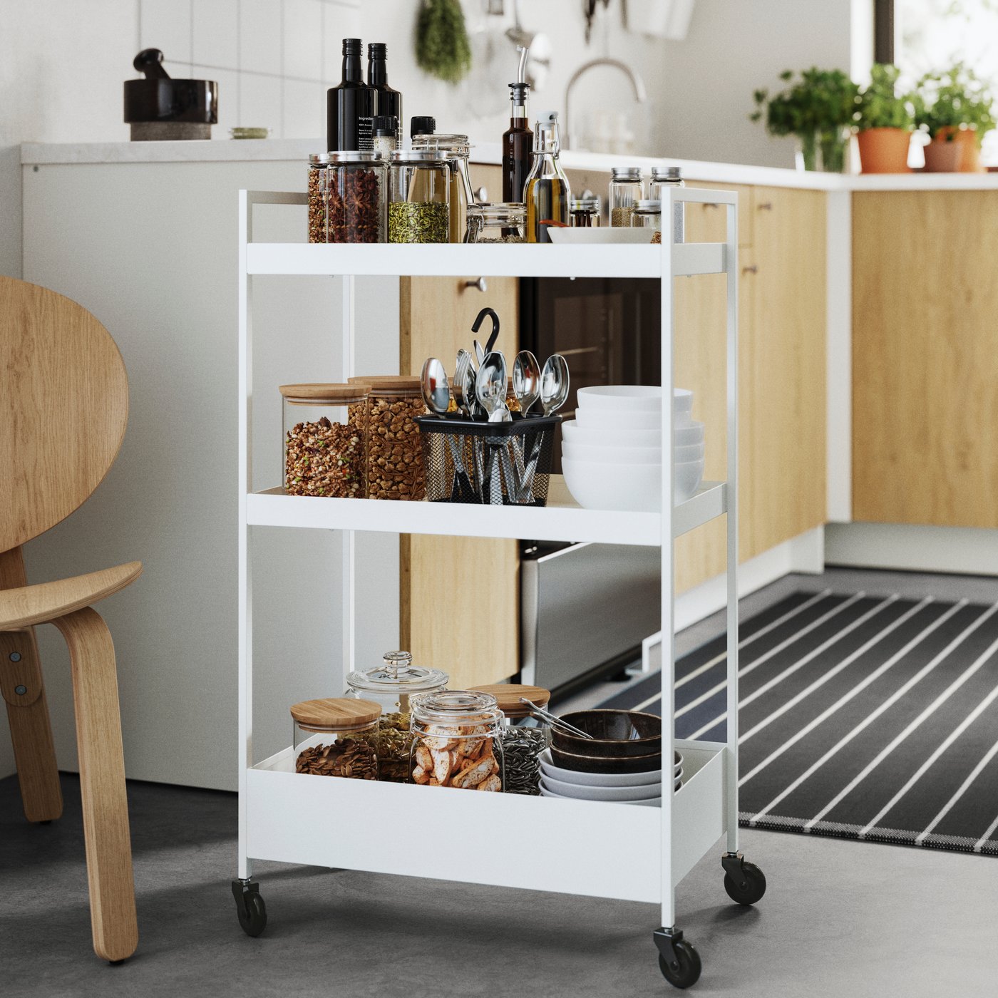 A white NISSAFORS trolley in a modern kitchen, filled with jars of spices, oils, and utensils on its three shelves.