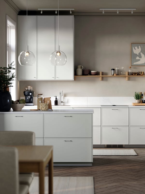 Helle, moderne IKEA Küche mit hell grauen ASPUDDEN Fronten, Kochinsel und dezenten Messingdetails; warmes, gemütliches Licht durch zwei Glaspendelleuchten, offene Wandregale mit Geschirr und Deko, im Vordergrund ein Holzesstisch – aufgeräumt und einladend für den Alltag.
