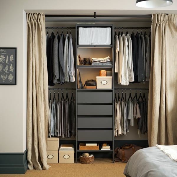 Clothes hang on the rails of a dark grey AURDAL wardrobe combination in a recess behind open beige curtains in a bedroom.