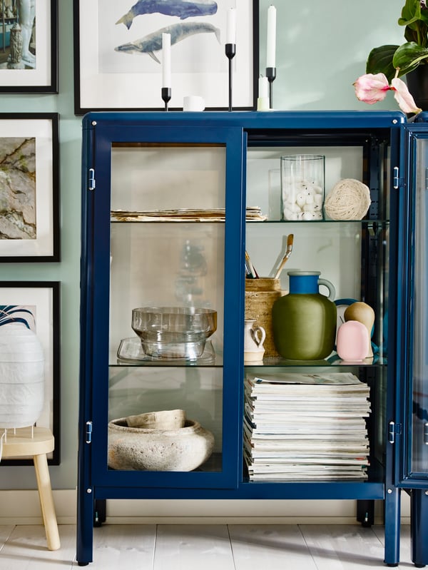 A FABRIKÖR glass-door cabinet in blue showing glassware, with candle sticks on top.