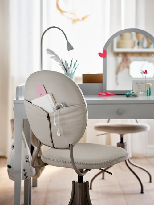 A grey desk with a BJÖRKBERGET chair in front of it. The chair's back pocket holds a book and a pair of headphones.