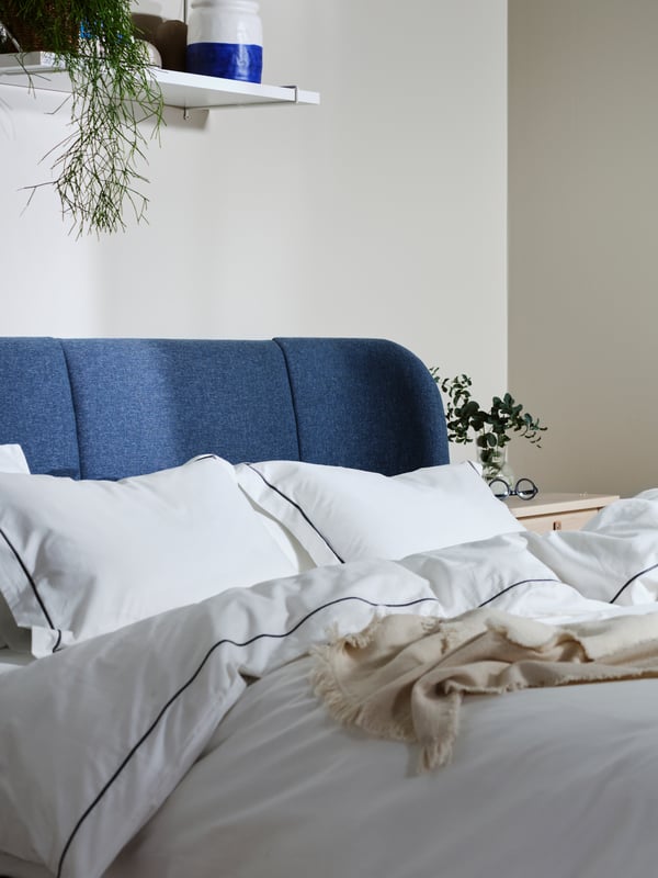 A white bedroom with an IDANÄS upholstered bed with no bed linen on, in front of two white IDANÄS wardrobes and a curtain.