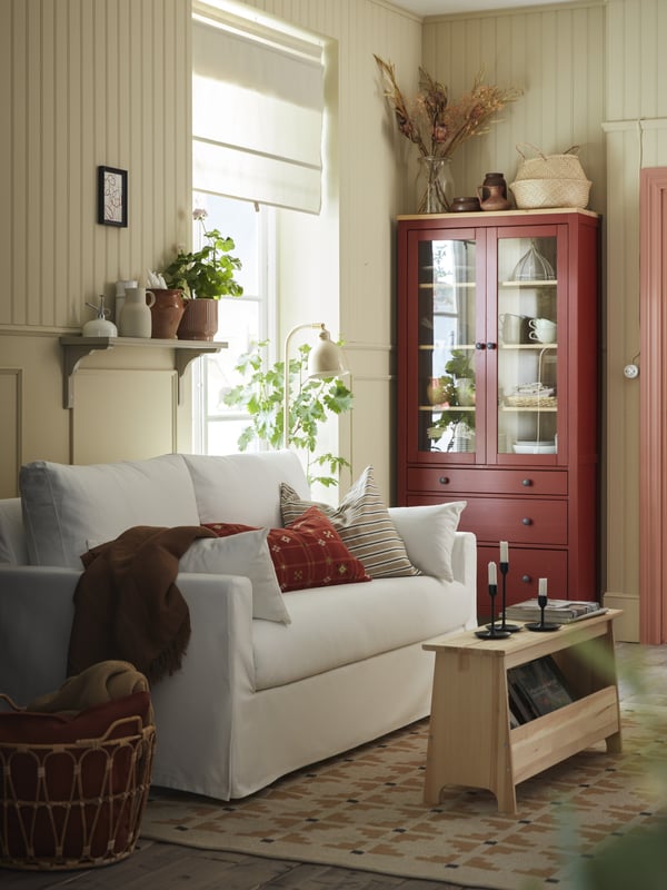 A white HYLTARP 2-seater sofa in the living room with a red HEMNES display cabinet and a colourful rug on the floor.