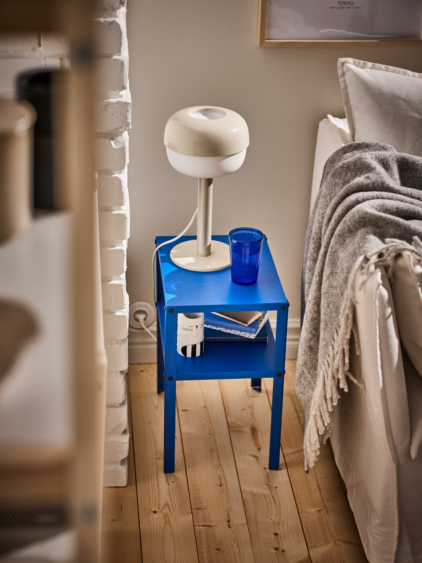 A dimly lit bedroom with a BLASVERK bedside table lamp sitting on a blue KNARREVIK side table. 