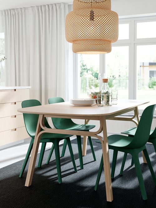 Dining room with a rectangular dining table with a carafe with stopper and green chairs, above which is a beige pendant lamp.