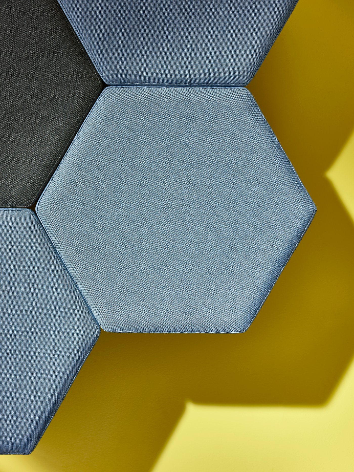 Four KJUGE pouffes, three in Knisa medium blue and one Knisa dark grey are placed together against a yellow backdrop.