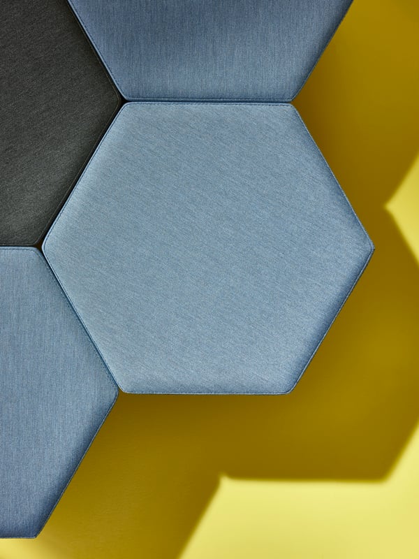 Four KJUGE poufs, three in Knisa medium blue and one Knisa dark grey are placed together against a yellow backdrop.