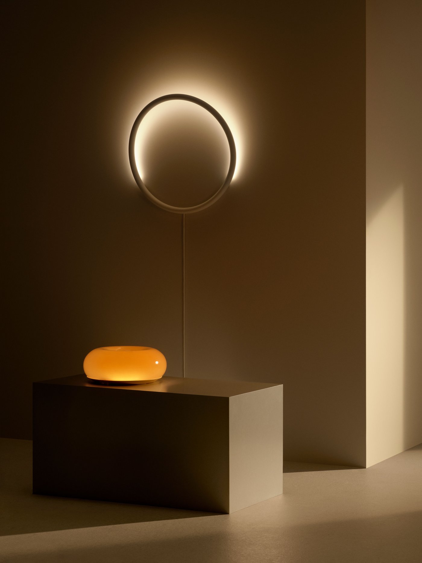 Minimalistiskt rum med VARMBLIXT vägglampa som lyser mjukt och ett lågt bord med en VARMBLIXT bord-/vägglampa, mot beige väggar och enkel belysning.
