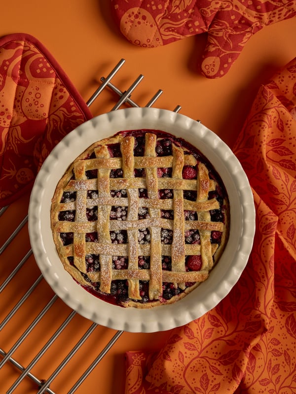 Torta ai frutti di bosco in una tortiera HÖSTAGILLE appoggiata su una superficie arancione, accanto a una presina, a un guanto da forno e a uno strofinaccio, anch'essi arancioni.