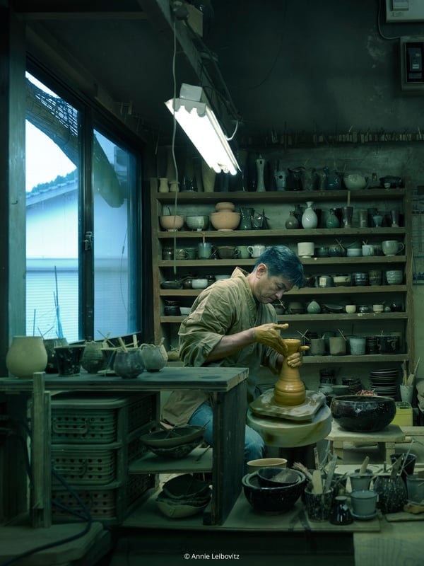 A man stands in a room filled with furnishings and tools and works with his ceramics.