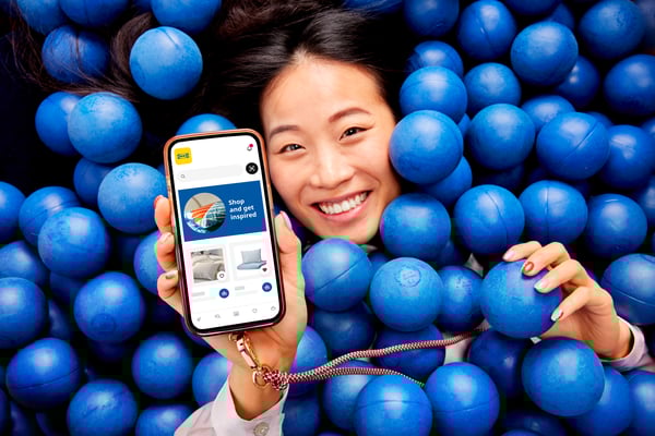 Uśmiechnięta kobieta zanurzona w basenie z niebieskimi piłkami trzyma w ręku smartfon z uruchomioną aplikacją IKEA widoczną na ekranie.