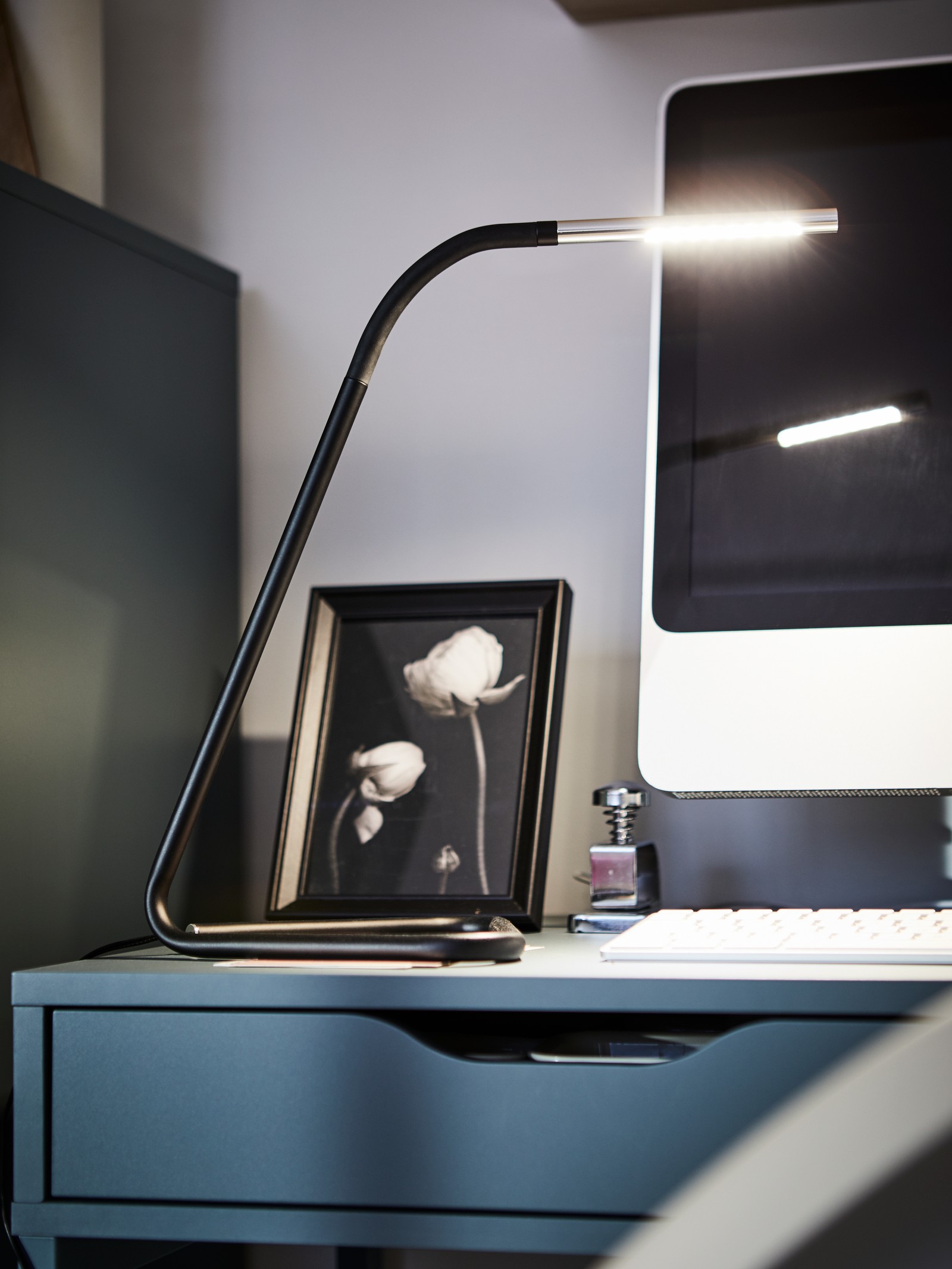 Una lámpara de trabajo LED HÅRTE negra sobre un escritorio azul e iluminada junto a la pantalla de un monitor y un teclado