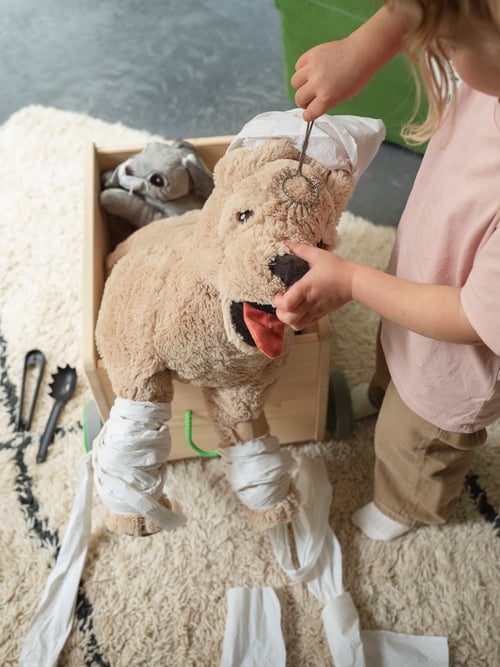Une jeune fille tient un fouet DUKTIG contre la tête d’une peluche GOSIG GOLDEN. Les pattes sont pansées avec du papier toilette.