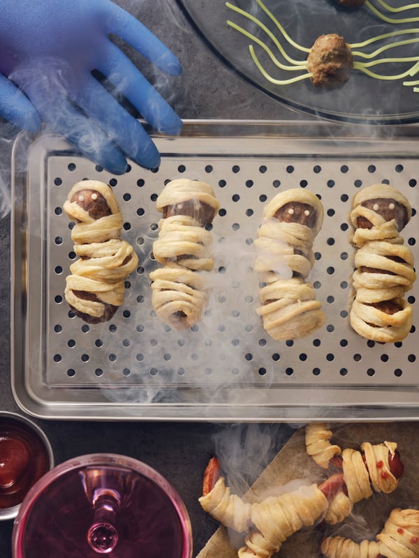 Quatre boulettes de viande HUVUDROLL enveloppées dans de la pâte pour ressembler à des momies sont disposées sur un plateau en acier, à côté d'autres plats sur le thème d'Halloween.