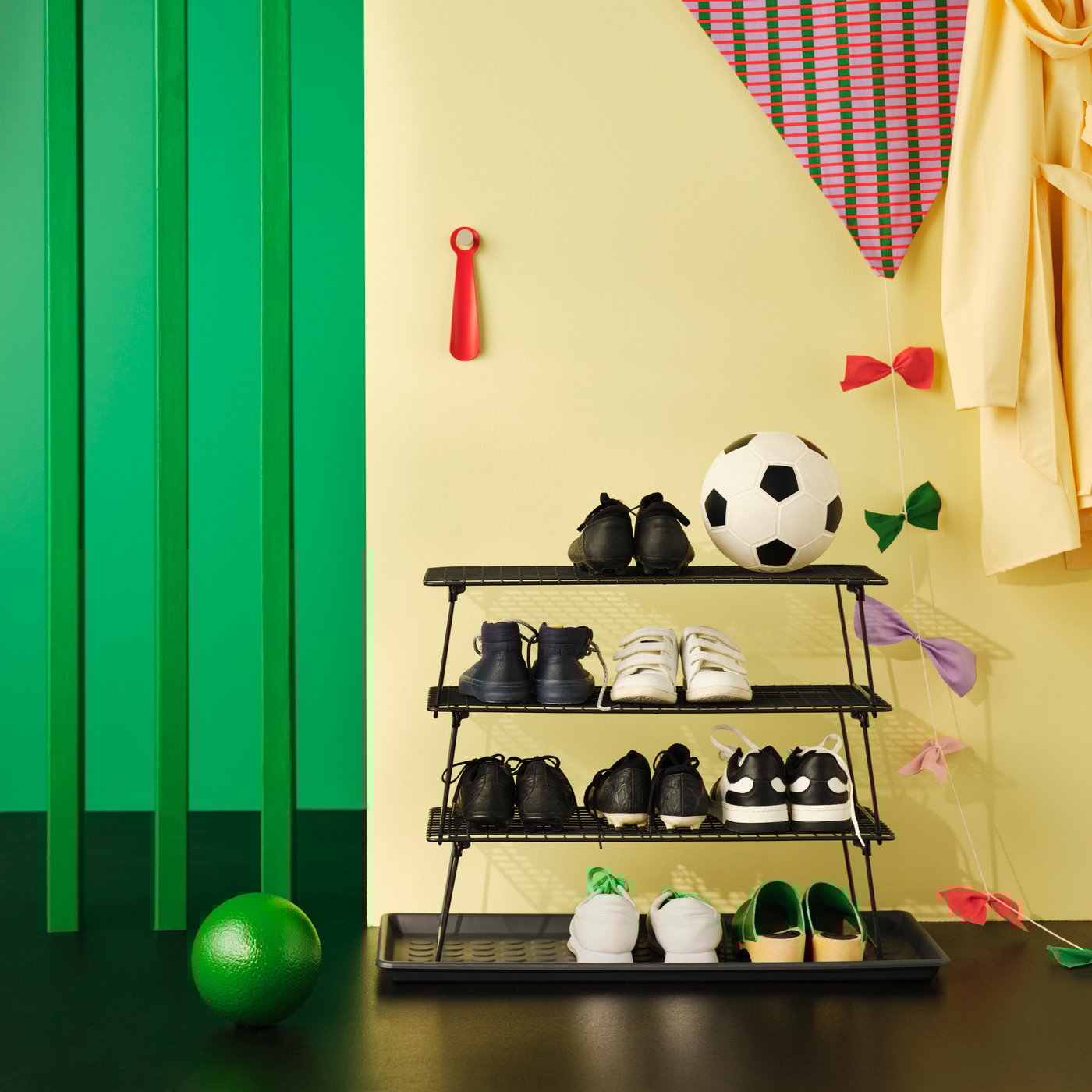 Trei suporturi de pantofi GREJIG stivuite pe o tavă pentru încălțăminte BAGGMUCK, pline cu încălțăminte sport și mingi, așezate lângă decorațiuni de perete pline de culoare.