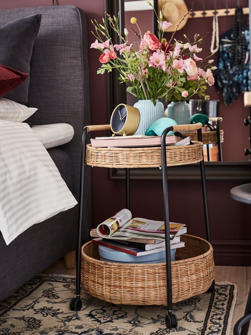 Bedroom with dark grey divan bed and black steel trolley table with two rattan baskets with vase of flowers and magazines.