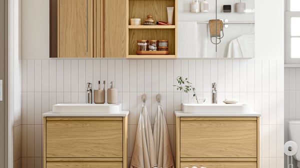 A white bathroom has ÄNGSJÖN bathroom furniture in oak effect, including two washstands with drawers and two mirror cabinets.