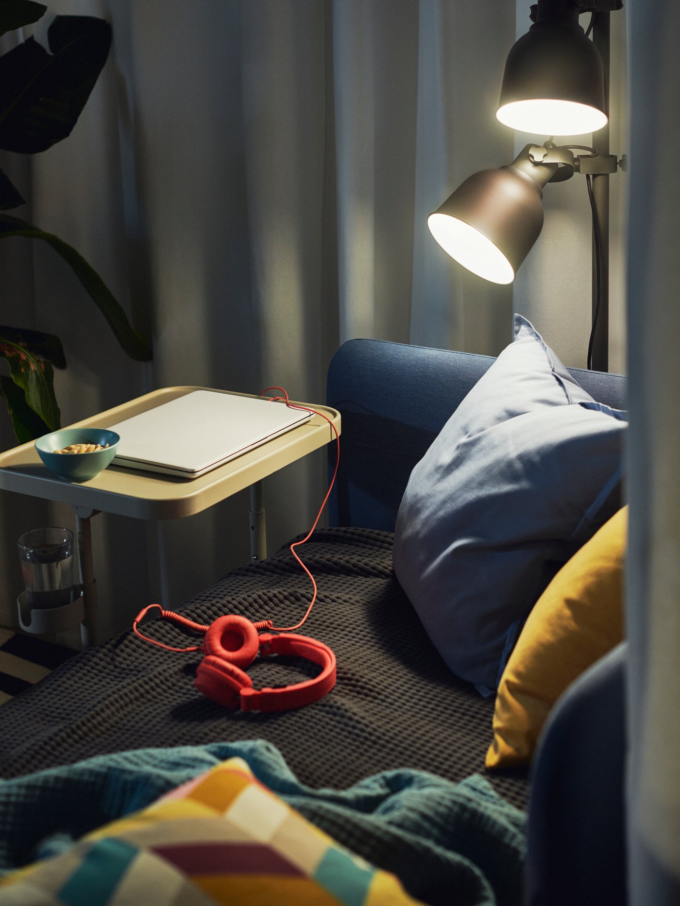 Una lámpara de pie HEKTAR gris oscuro ilumina una cama BLÅKULLEN cubierta de textiles y con un soporte para ordenador portátil BJÖRKÅSEN junto a ella.