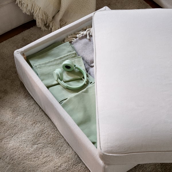 A light living room with a partly open KIVIK footstool with textiles stored inside, placed on a beige STOENSE rug.