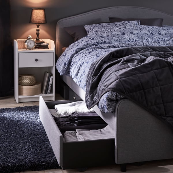 A grey HAUGA bed with a grey storage box pulled out underneath filled with bedlinen. A white bedside table with a lamp.