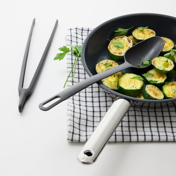 A FULLÄNDAD cooking spoon is lying in a frying pan with zucchini. Next to the pan, a pair of FULLÄNDAD cooking tweezers.
