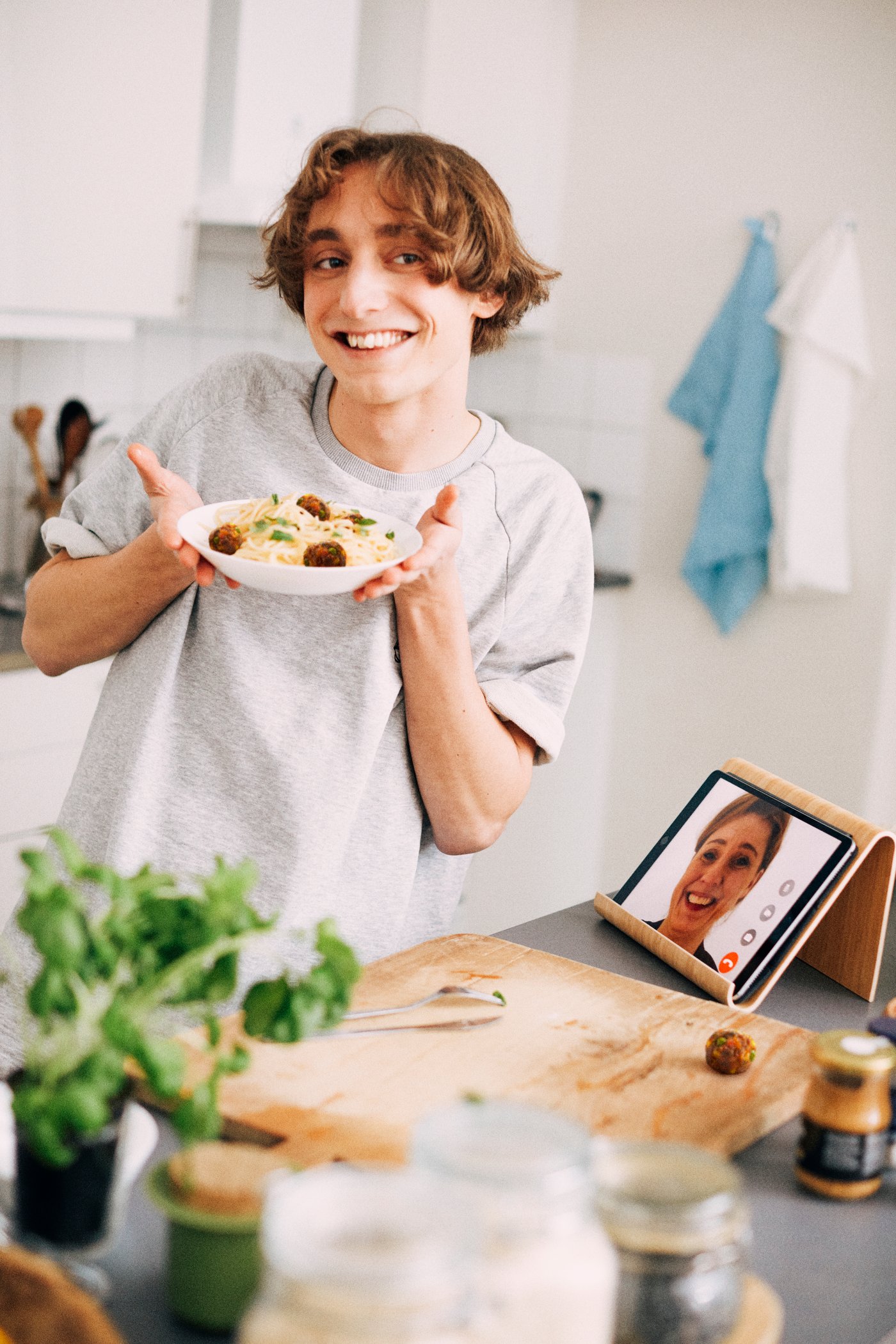 A person in a kitchen holds up a plate of food while video calling someone on a tablet that’s propped up on the counter. Fresh herbs, jars, and cooking ingredients are spread out on the workspace.