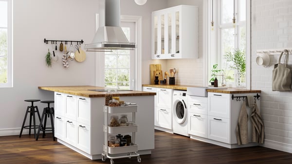 An IKEA AXSTAD white shaker kitchen with glass doors, oak worktop and black door knobs. A kitchen island made of the same kitchen drawers and a white trolley on the side.