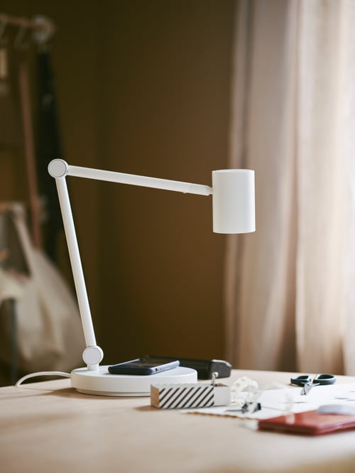 Un prim-plan al unei lămpi de lucru albe cu încărcare wireless, o pereche de foarfece și o cutie, toate așezate pe o suprafață bej.