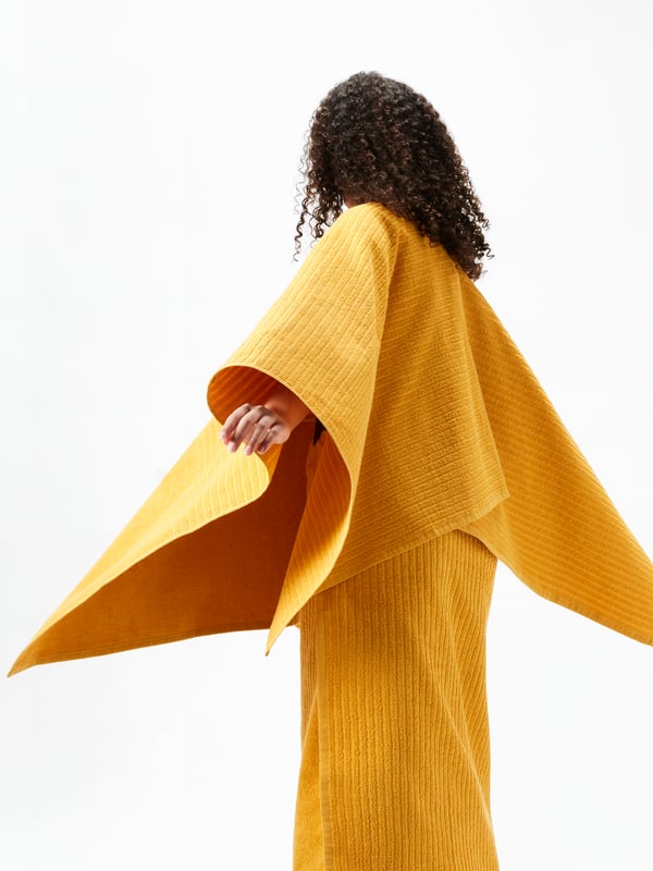 A side view of a person with their arm stretched as if in mid twirl, wearing three yellow VÅGSJÖN towels like clothing.