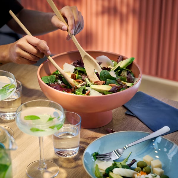 A person is using wooden utensils to take some salad from a red-brown BLOMVECKLARE serving bowl that’s placed on a table.
