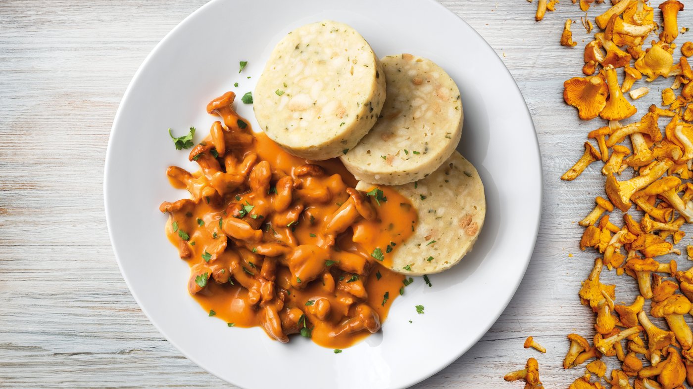 Auf einem weißen Teller liegen in Scheiben geschnittene Knödel mit einer cremigen Eierschwammerl Sauce. Neben dem Teller sind weitere Eierschwammerl dekorativ auf einer hellen Holzoberfläche verteilt.
