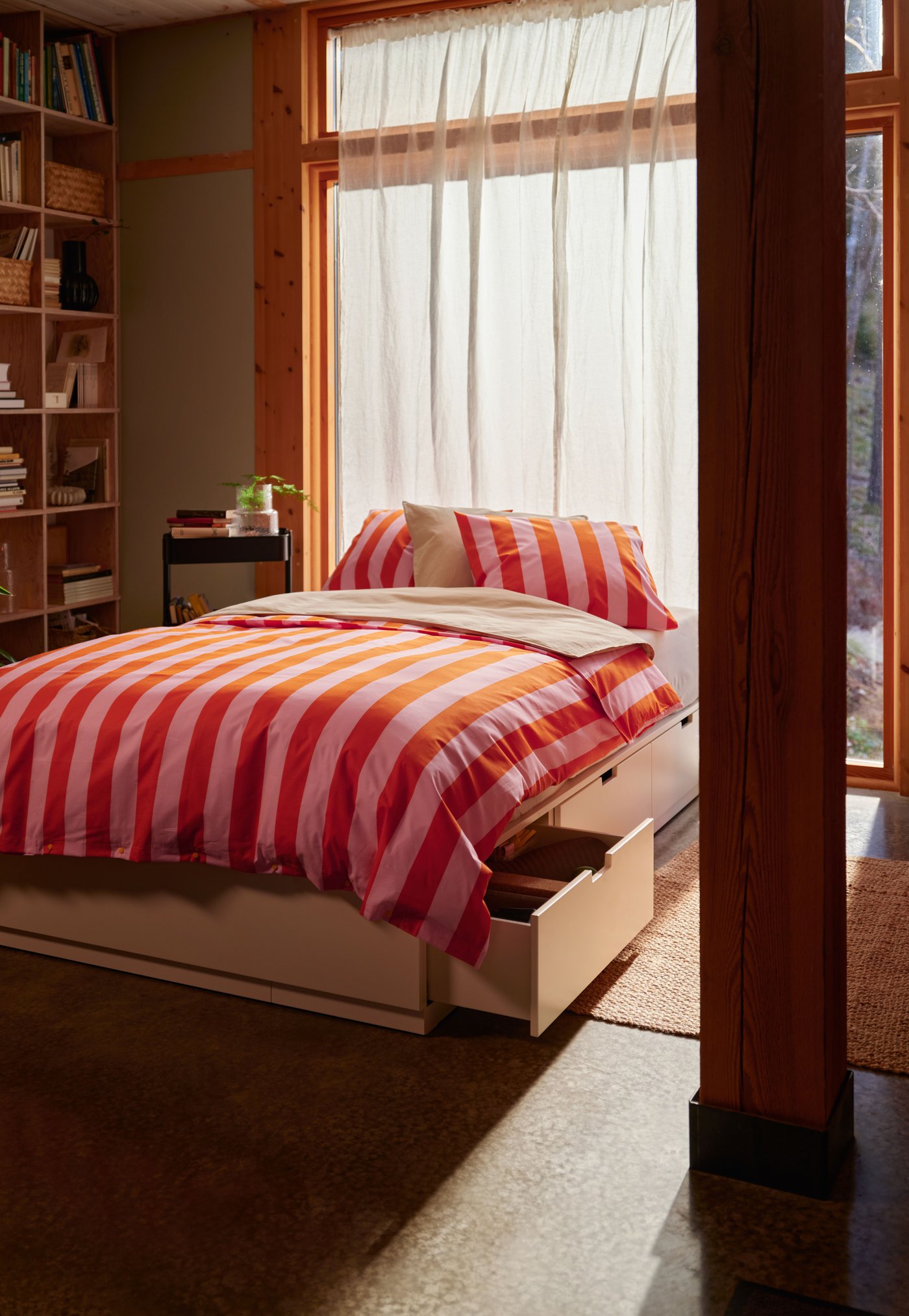 NORDLI äng med röd‑ och rosarandigt bäddset i ett träfyllt rum, med utdragen förvaringslåda och stort fönster med tunna gardiner som släpper in ljus.