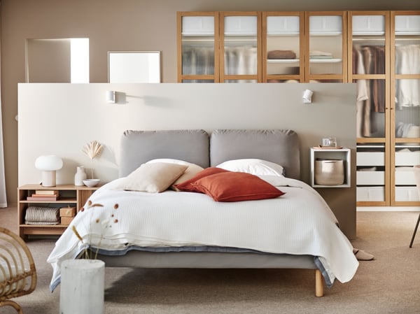 A SAGESUND upholstered bed in Diseröd brown/Luröy in a large airy bedroom covered in white bed linen with red cushions on it.
