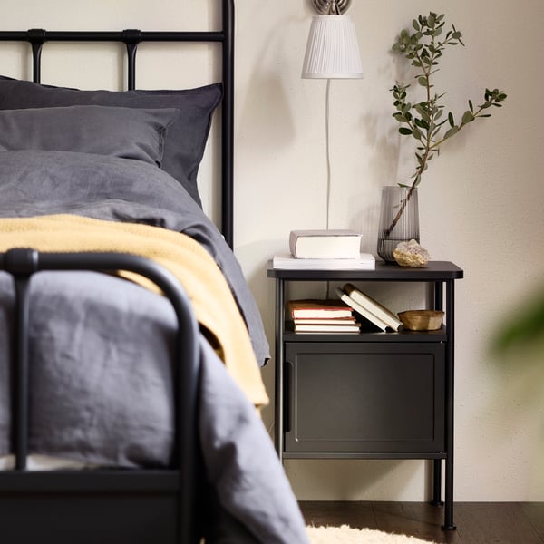 GRÅFJÄLLET bed side table propped with books and stuff, standing next to STJÄRNÖ bed frame with dark grey bedding.