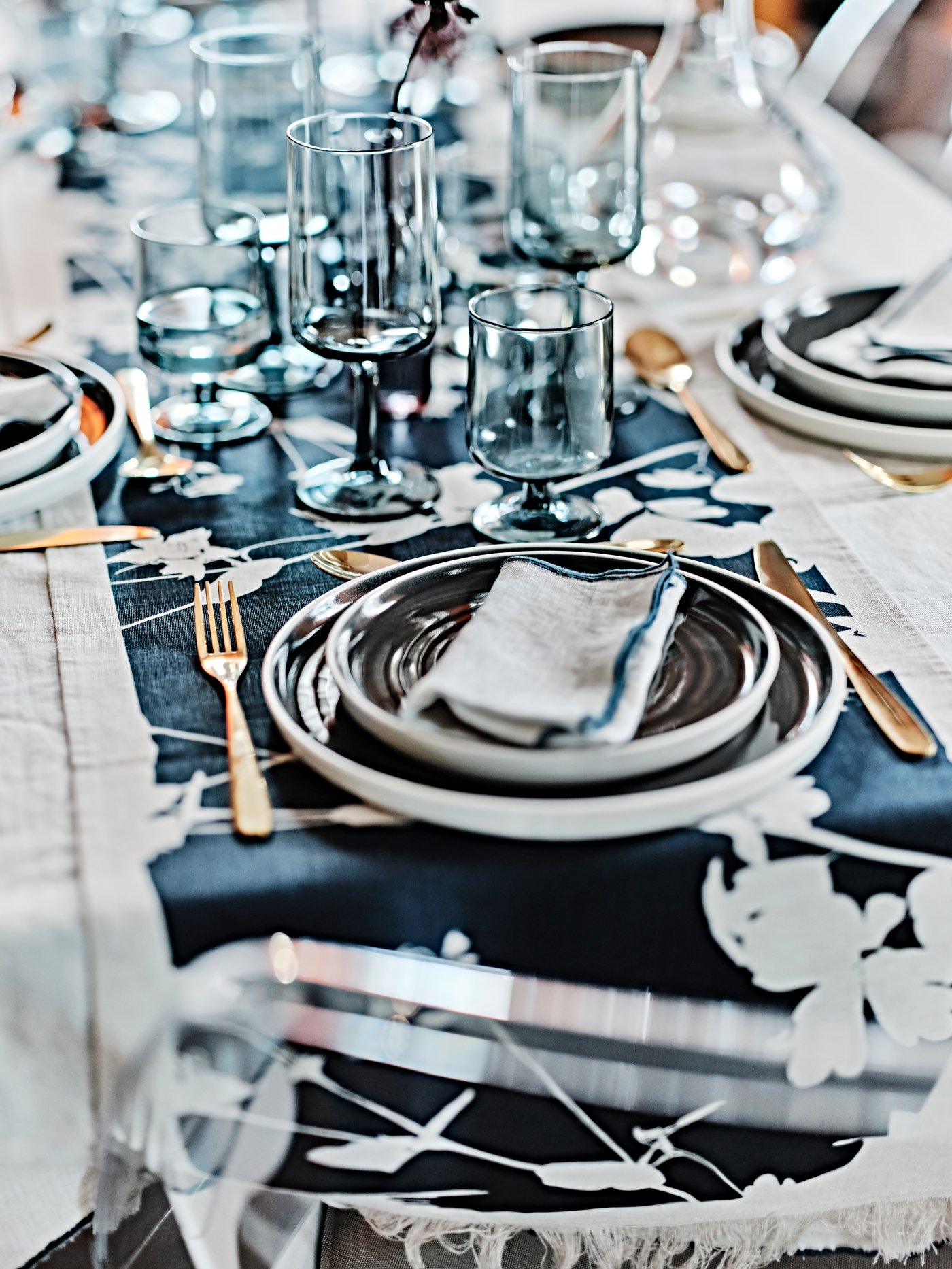 A table laid with a brass-colour TILLAGD 24-piece cutlery set, dark grey OMBONAD side plates and plates and OMBONAD glasses.