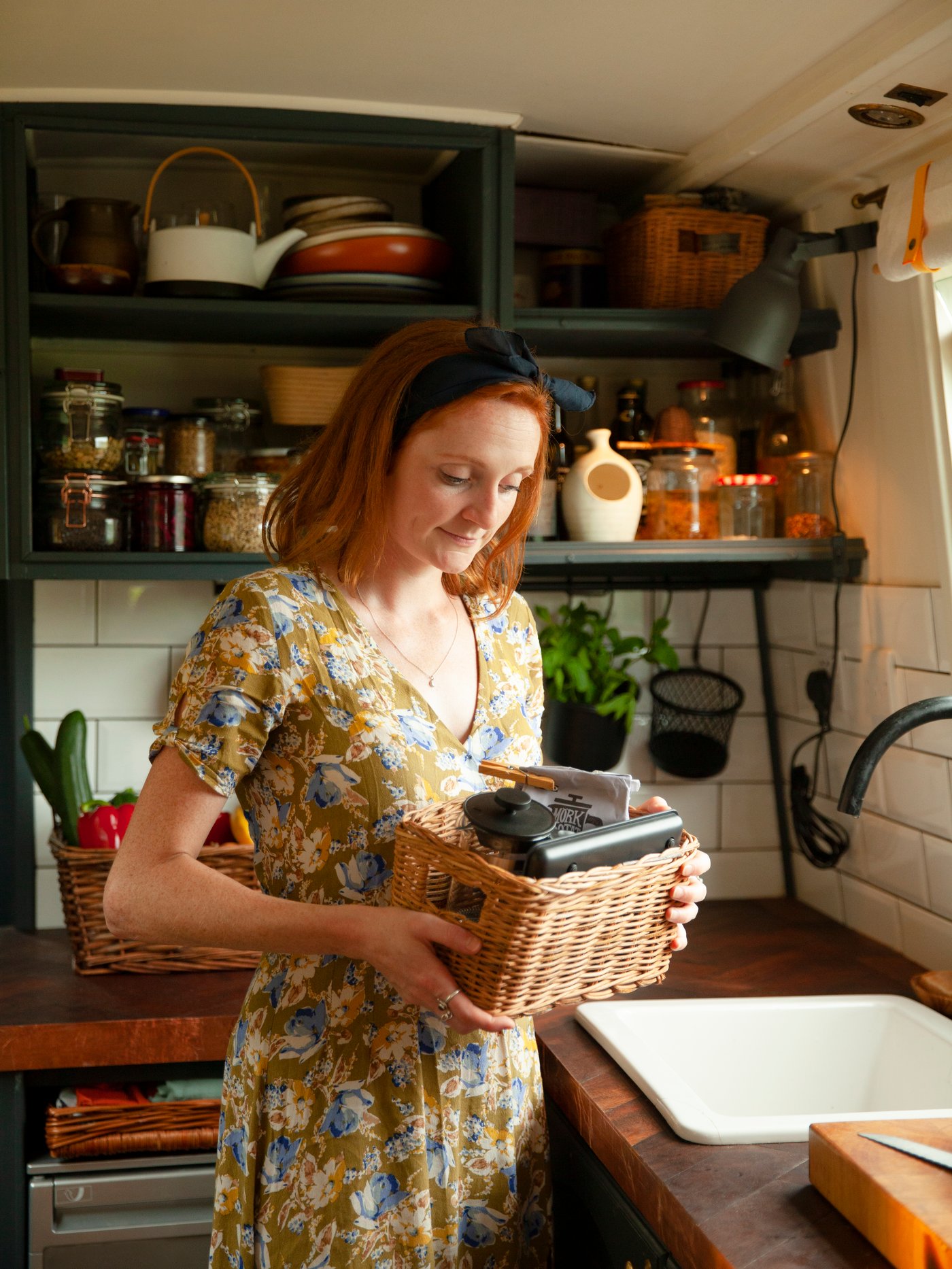 Une femme dans une cuisine porte un petit panier contenant son café PÅTÅR, sa cafetière UPHETTA et une balance électrique GENOMSITT.